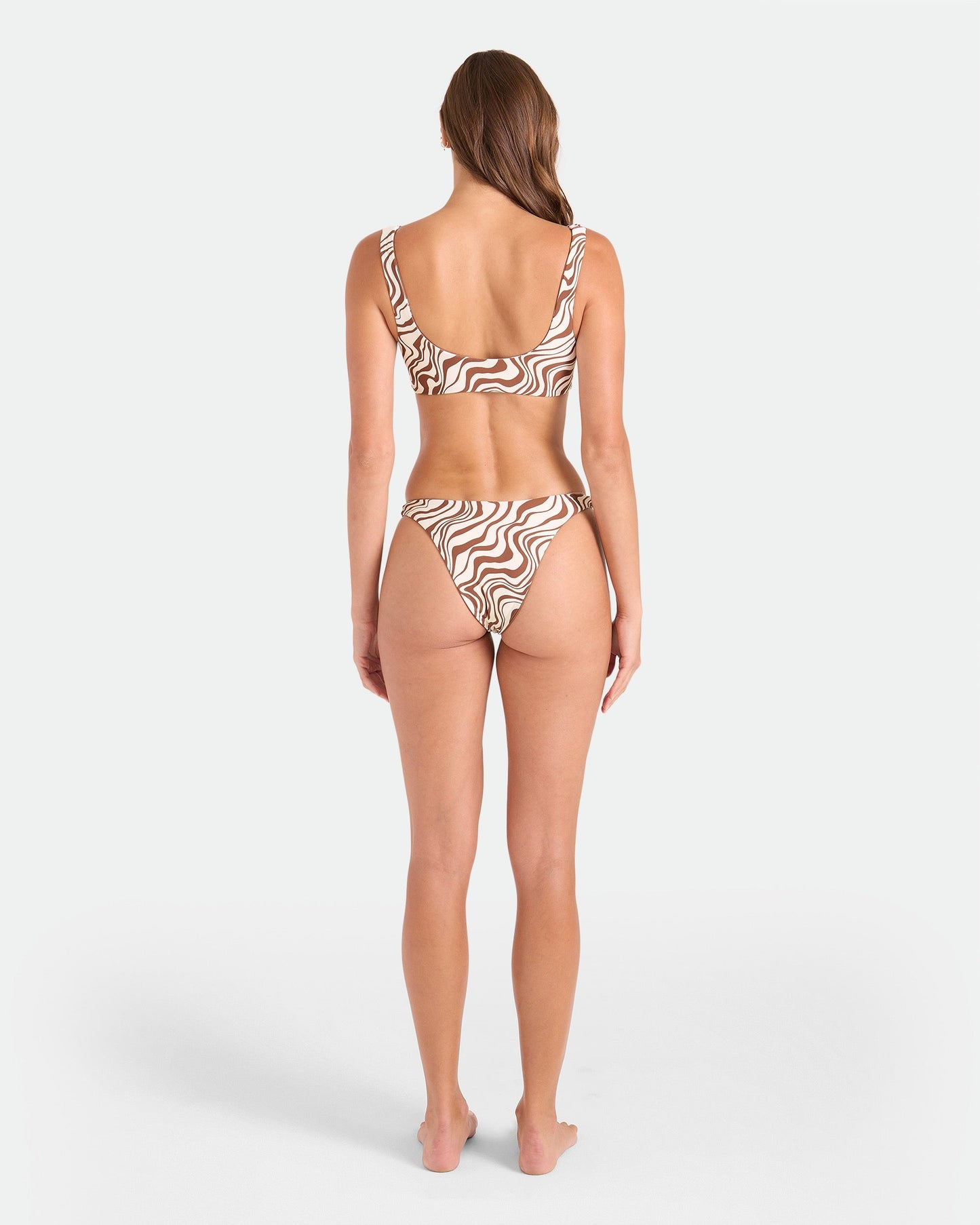 A woman stands barefoot facing away from the camera wearing the Volcom Women's Coco Retro Bloom Hipster Cheeky Reversible Bikini Bottom, featuring a brown and white abstract pattern, with her long hair swept to one side against a plain white background.