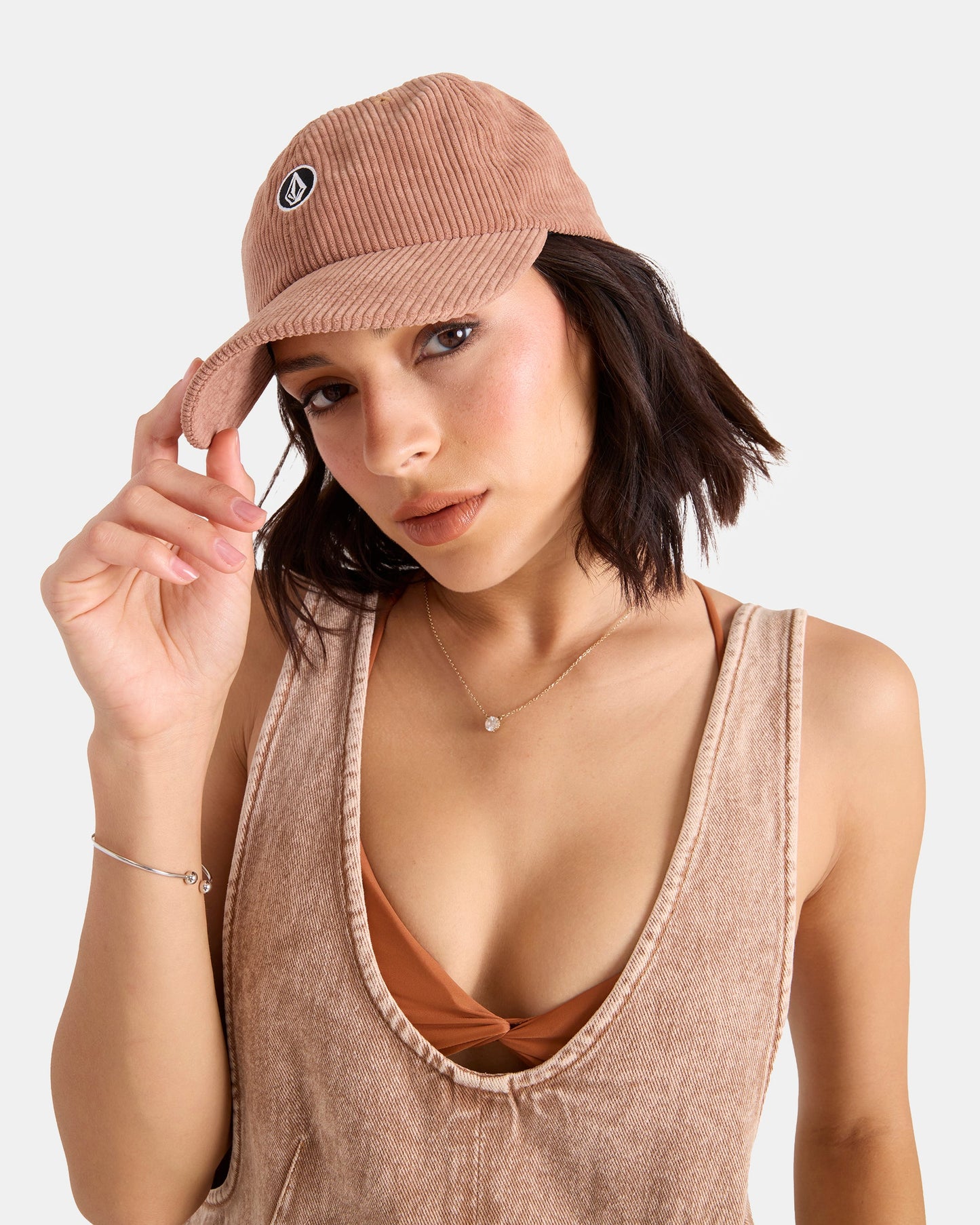 A woman with short dark hair wears the Volcom Women's Circle Stone Cord Hat and a matching sleeveless top, touching the brim of her cap as she looks at the camera in front of a plain white background.