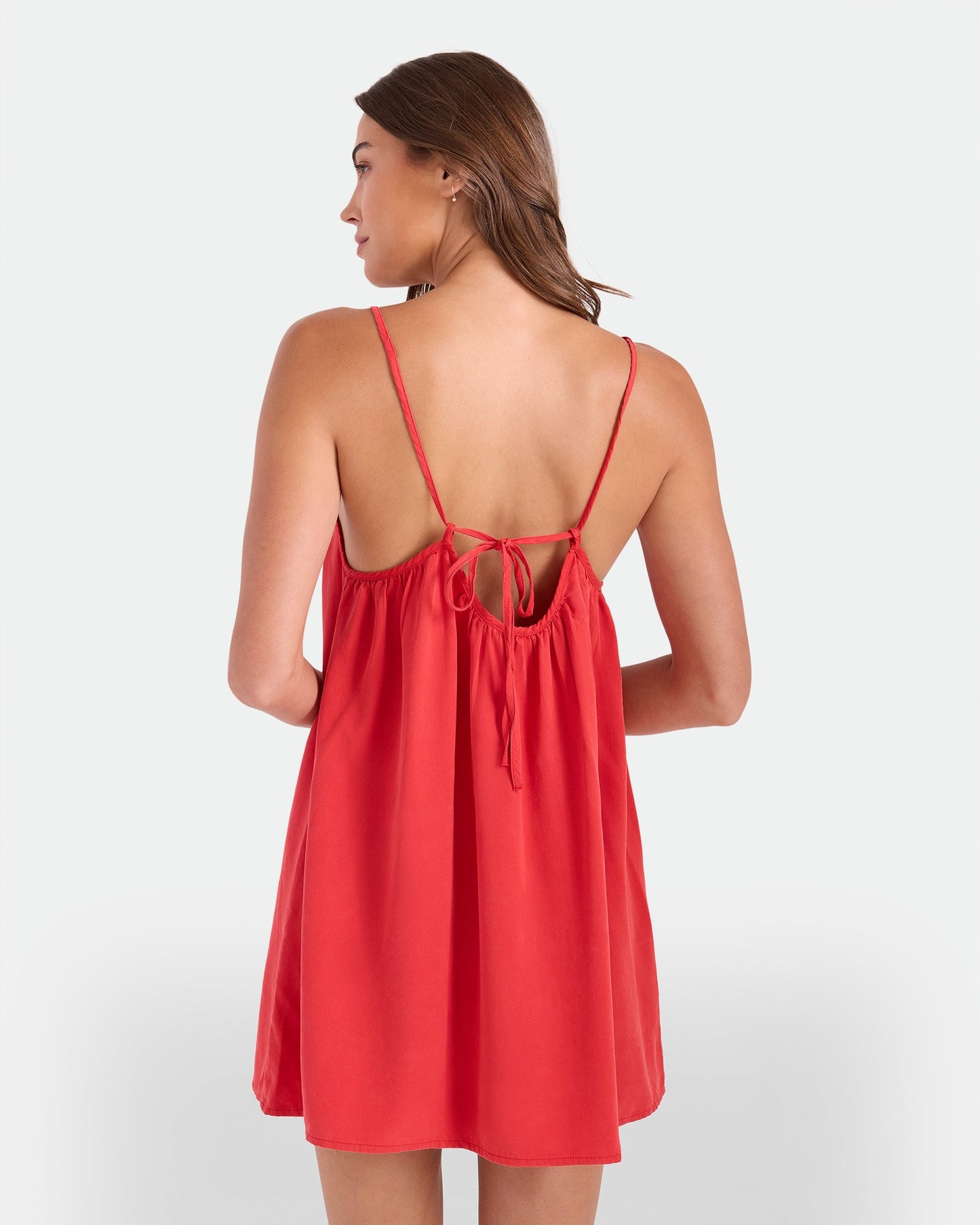 A woman with long brown hair wears the Volcom Women's Baby Cakes Mini—a loose red, sleeveless dress with thin straps and an open back tied with a bow—standing facing away from the camera against a plain background.