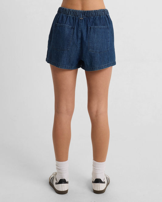 A woman faces away, wearing the Women's Stone Shine Short by Volcom in dark blue denim with side and back pockets, paired with white socks and white sneakers featuring black accents. The background is plain white.