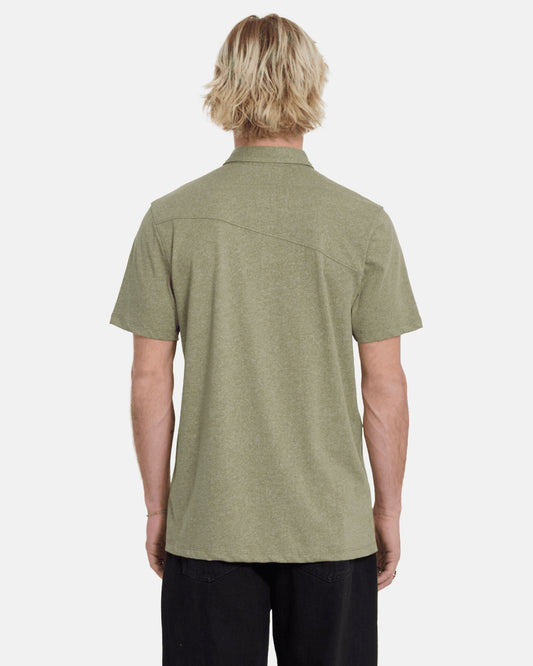 A person with light blonde, wavy hair is seen from behind wearing the Volcom Men's Wowzer Polo Short Sleeve Shirt in olive green and black pants against a plain, light background.