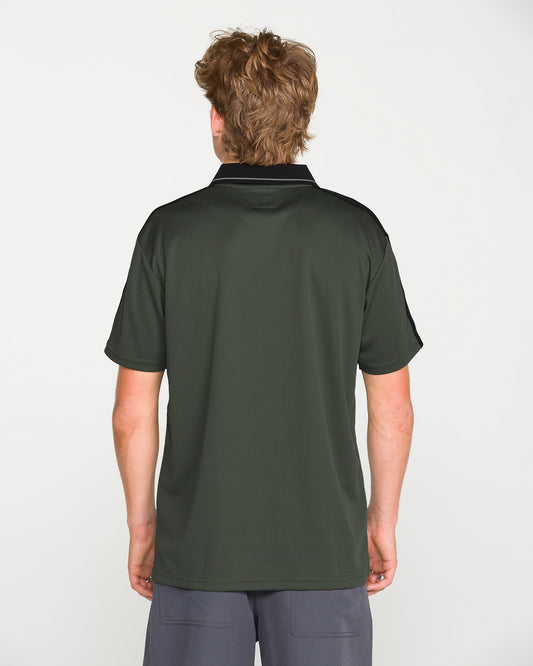 A person with light brown hair stands facing away, wearing the Volcom Men's Chillstone Short Sleeve Polo in dark green with a black collar and shoulders featuring subtle yarn-dye stripes, paired with gray shorts, against a plain white background.