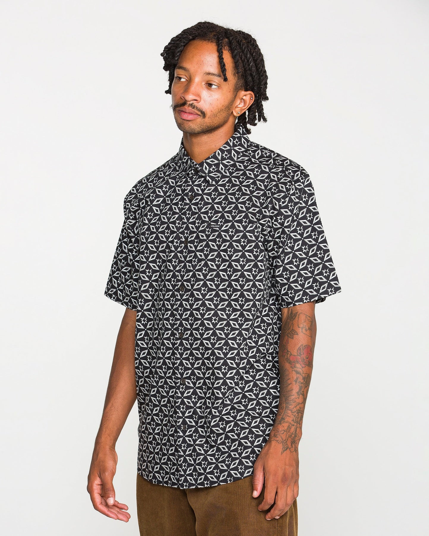 A man with short dreadlocks and tattoos on his arm wears the Volcom Men's Scaler Stone Short Sleeve Shirt and brown pants, standing against a plain white background.