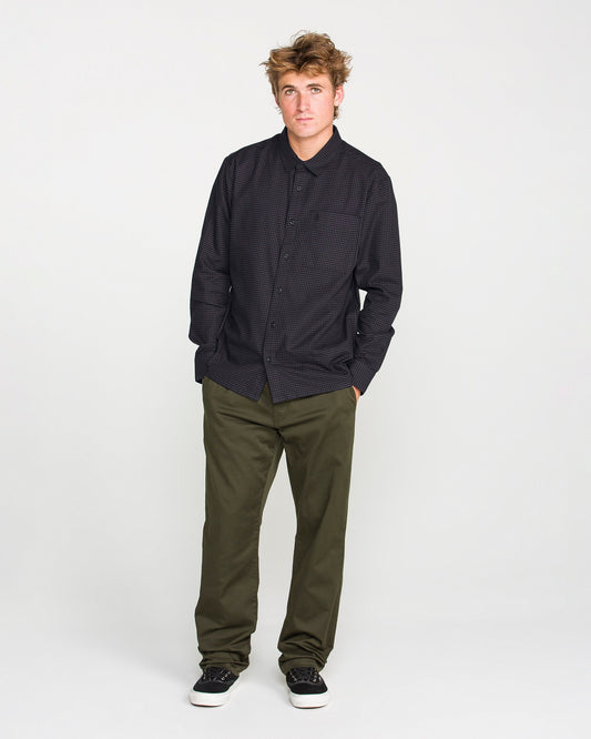 A young man with tousled hair stands against a plain white background, wearing the Volcom Men's Glasnow Long Sleeve Shirt in a dark, boxy fit with a yarn-dyed dobby texture, paired with olive green pants and black sneakers with white soles. His hands are in his pockets as he looks slightly to the side.