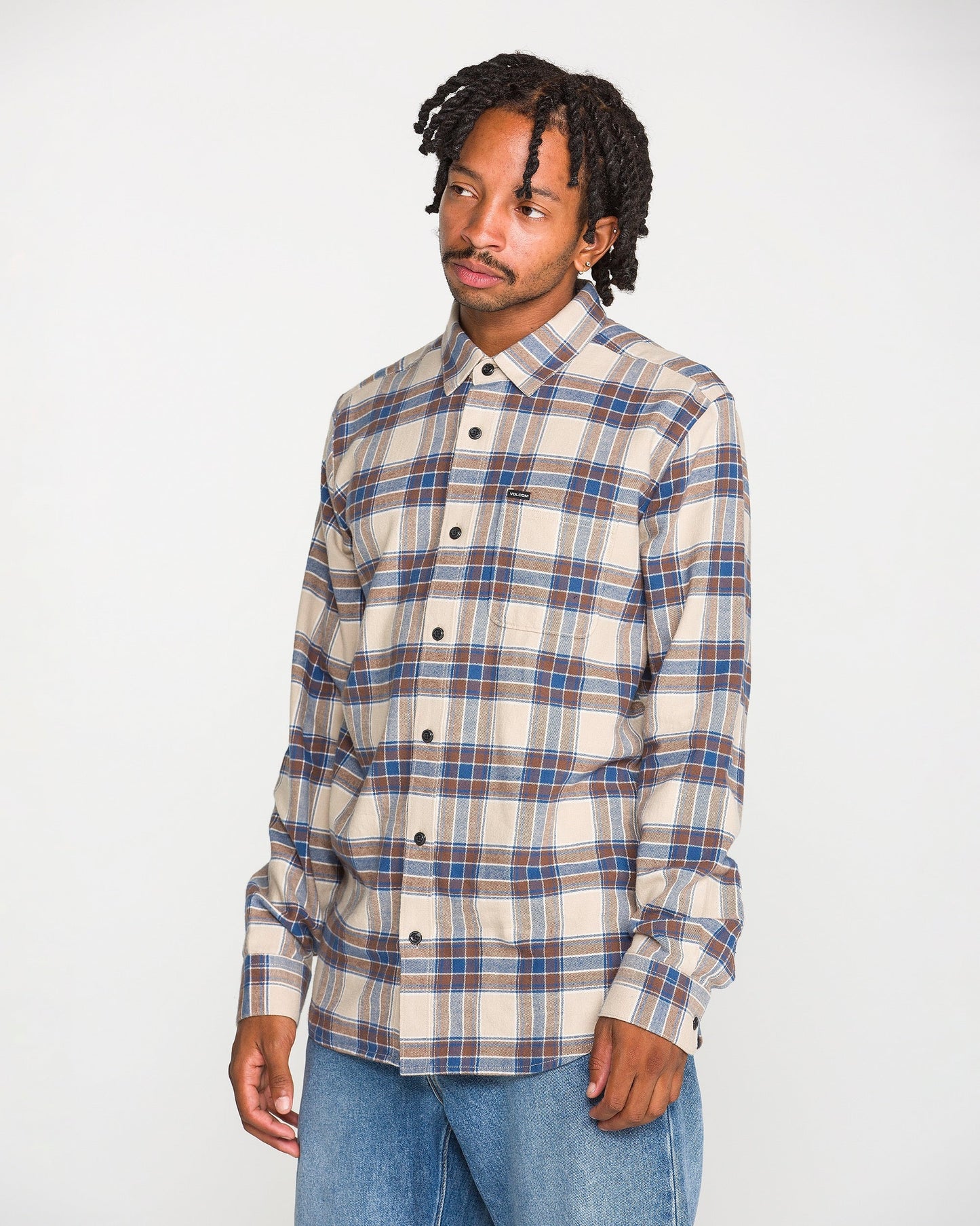 A man with short dreadlocks wears the Volcom Men's Caden Plaid Long Sleeve Shirt, featuring a classic fit in beige, blue, and brown plaid, paired with light blue jeans. Crafted from soft cotton flannel, the shirt stands out as he poses against a plain white background, looking slightly to his left with a neutral expression.