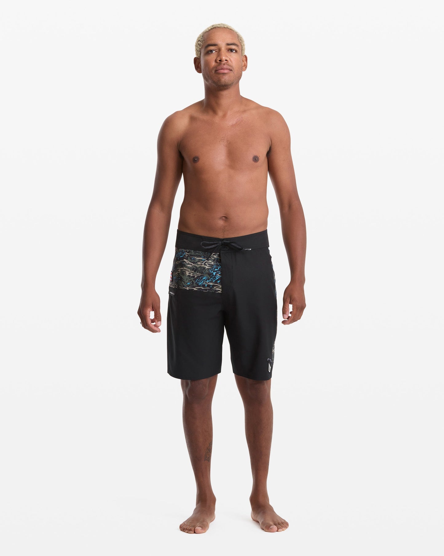 A man with short, bleached hair stands barefoot against a white background, wearing Volcom Men's Parillo Mod 21 black board shorts with a patterned side. He is shirtless, facing forward with a neutral expression.