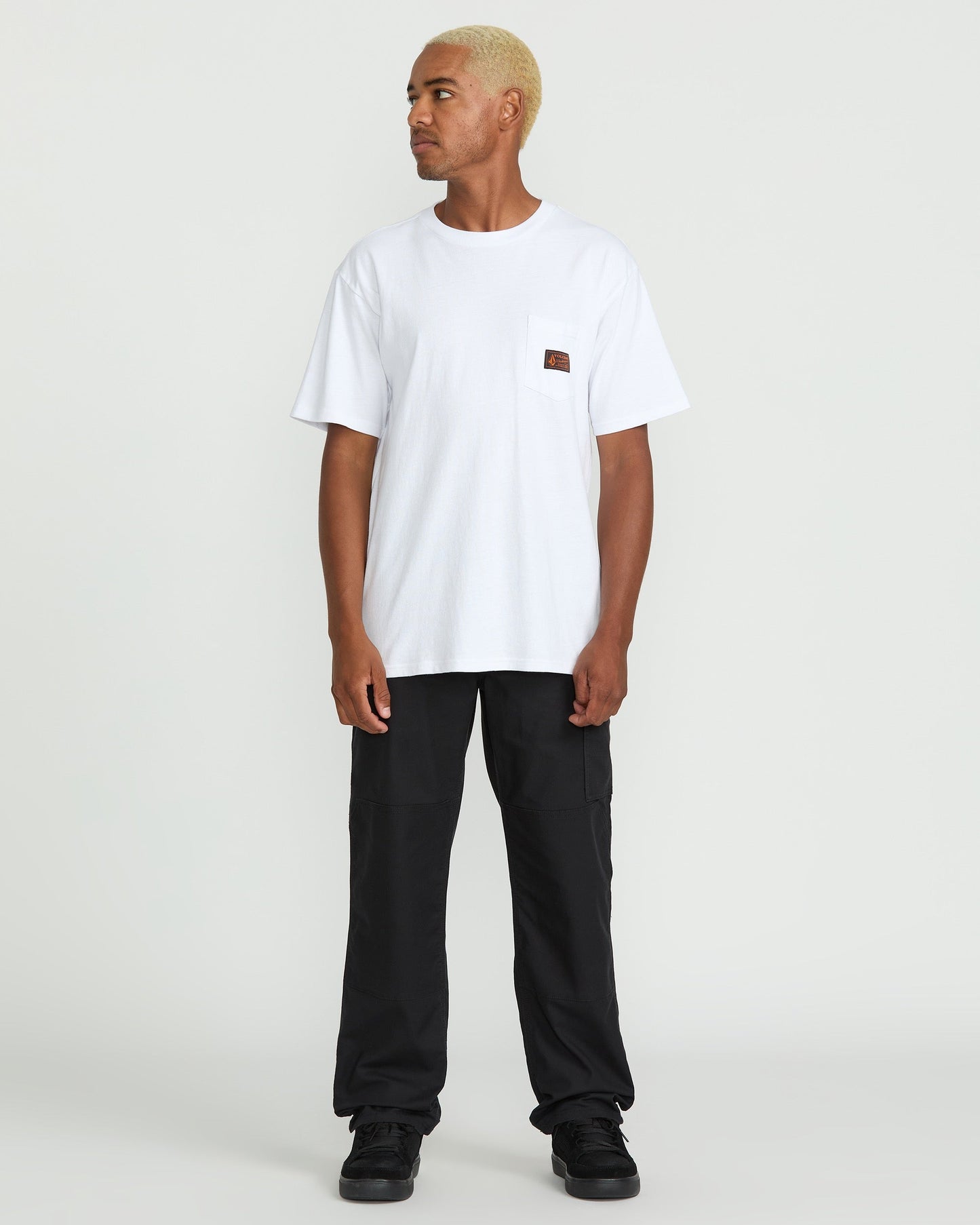 A person with short blond hair stands slightly to the side, wearing a white pocket t-shirt, Volcom Men's Caliper II Relaxed Work Pants in black stretch canvas with cargo pockets, and black sneakers against a plain white background.