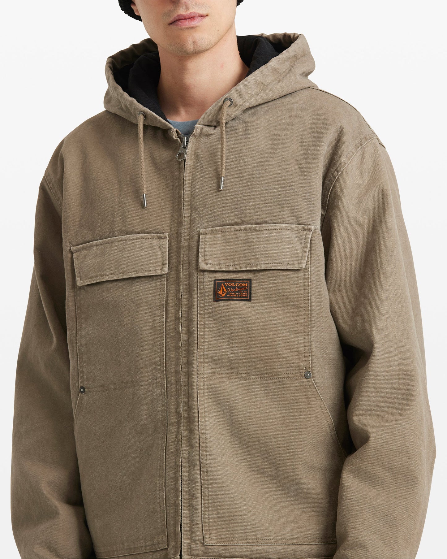 A person wears the Volcom Men's Freshdust Workwear Jacket, featuring large hand and chest pockets, a zip front, and a small rectangular patch on the left chest. Their face is partly visible against a plain background.