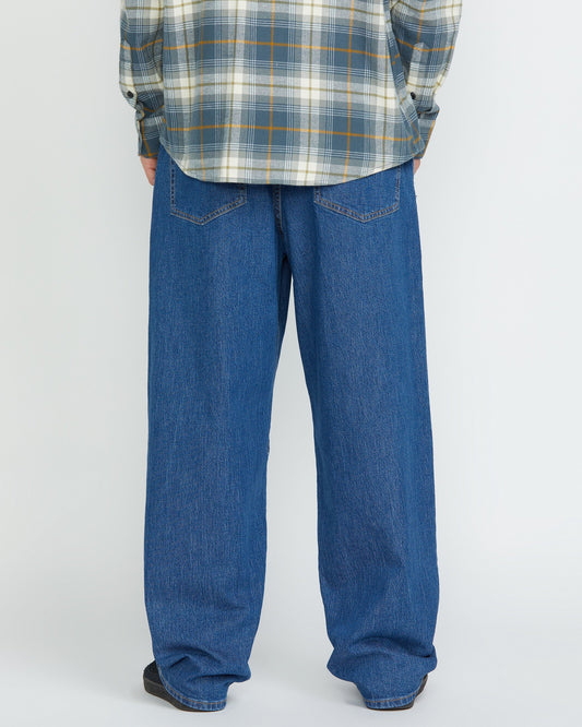A person stands facing away, showing only the lower half, wearing Volcom Men's Billow Denim Pants and a gray plaid shirt against a plain light background.
