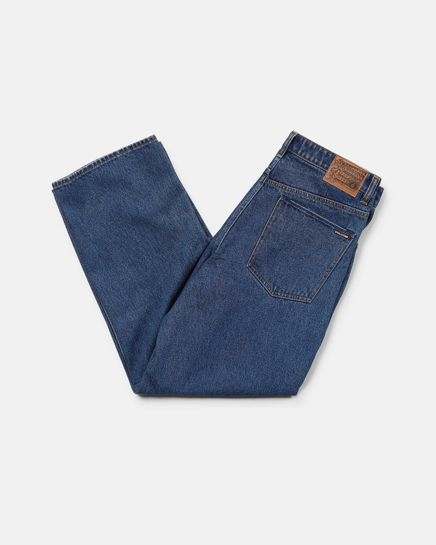 A pair of Volcom Men's Billow Denim Pants in dark indigo is neatly folded, showcasing a brown leather patch on the waistband and a visible back pocket. The low rise jeans are displayed against a plain white background.