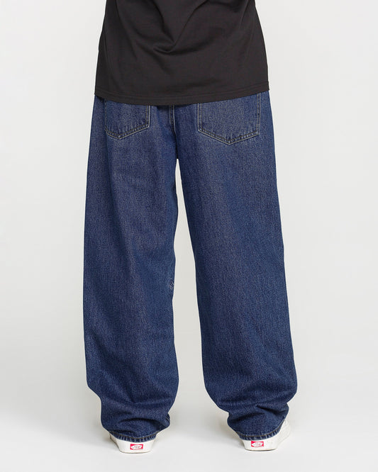 A person is standing against a plain light background, photographed from behind to show only the lower half of their body. They are wearing Volcom Men's Billow Denim Pants in a dark indigo wash, paired with a black shirt and white sneakers.