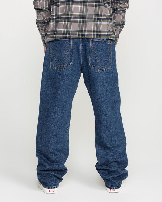 A person wearing a plaid shirt and Volcom Men's Pipe Dreams Denim Pants in a dark indigo wash stands facing away, showcasing the back of the jeans and white sneakers against a plain background.