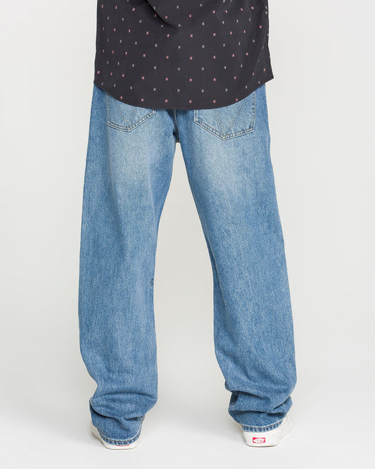 A person wearing Volcom Men's Pipe Dreams Denim Pants in Euro Light Blue, paired with a black shirt featuring a subtle pattern and white sneakers, stands with their back to the camera.