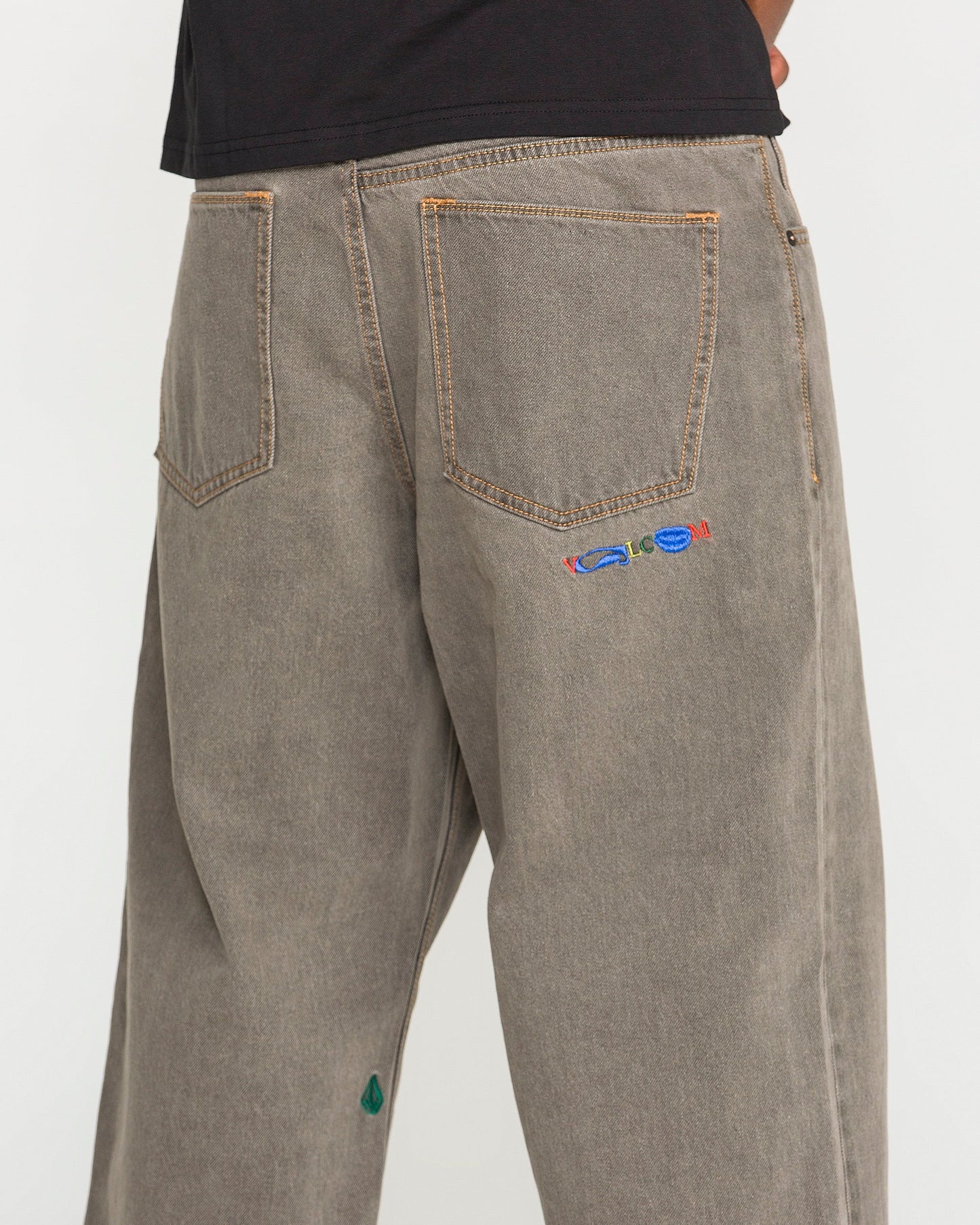 A person is seen wearing the Volcom Men's Featured Artist Antonio Carrau Reverb Pants—loose, gray-toned jeans with visible orange stitching, a vibrant embroidered motif near the right back pocket, and a small green accent by the left hem. Their upper body is partly shown in a black shirt.