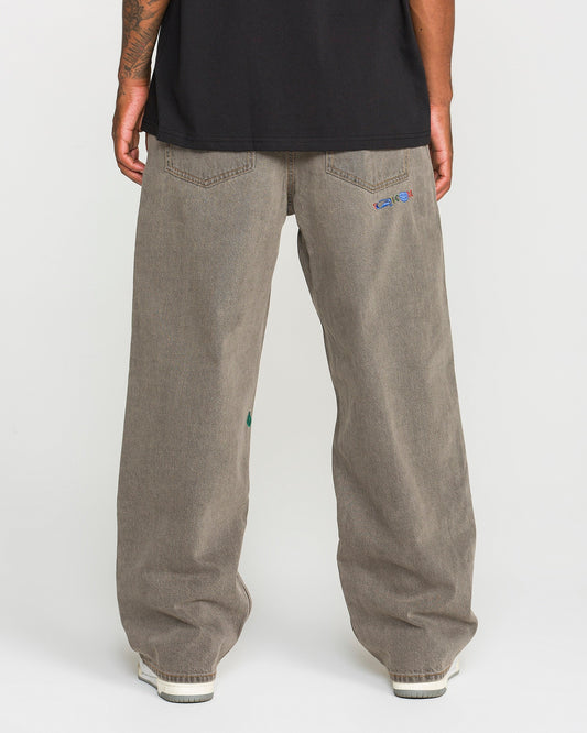 A person is standing with their back to the camera, wearing Volcom Men’s Featured Artist Antonio Carrau Reverb Pants—loose-fitting, low-rise gray jeans with small embroidered details on the back pockets—and a black shirt made of overdyed cotton. White shoes are partially visible.