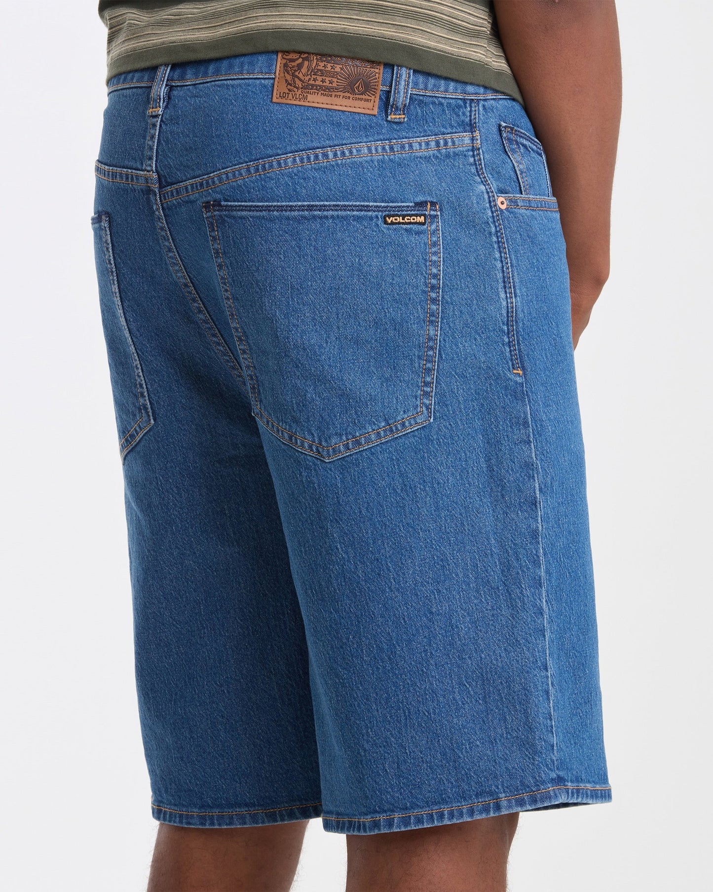 Someone wearing Volcom Men's Billow Denim Shorts featuring back pockets, a brown patch label near the waistband, and a visible Volcom logo on one pocket, paired with a green striped shirt.