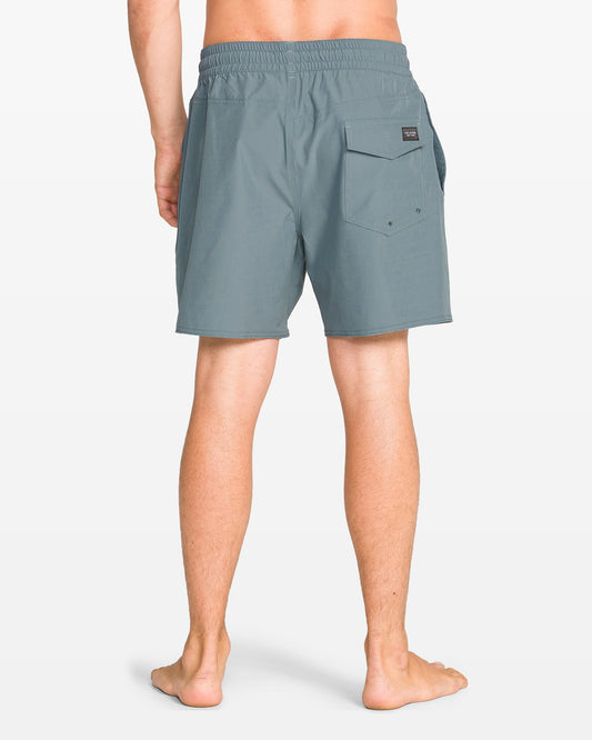 A person stands barefoot facing away, wearing Volcom Men's Lido Solid 16" Trunk boardshorts in teal with a 5-inch inseam, elastic waist, right flap pocket, and a small black label above the pocket. Background is plain white.