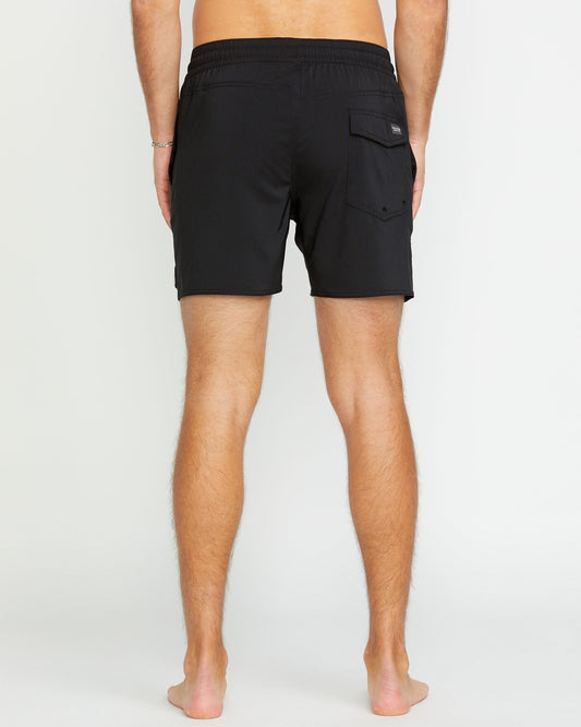 A barefoot person stands with their back to the camera, wearing Volcom Men's Lido 2 Solid Trunks—short black swim trunks made from recycled polyester, featuring an elastic waistband and a single flap pocket on the right side.