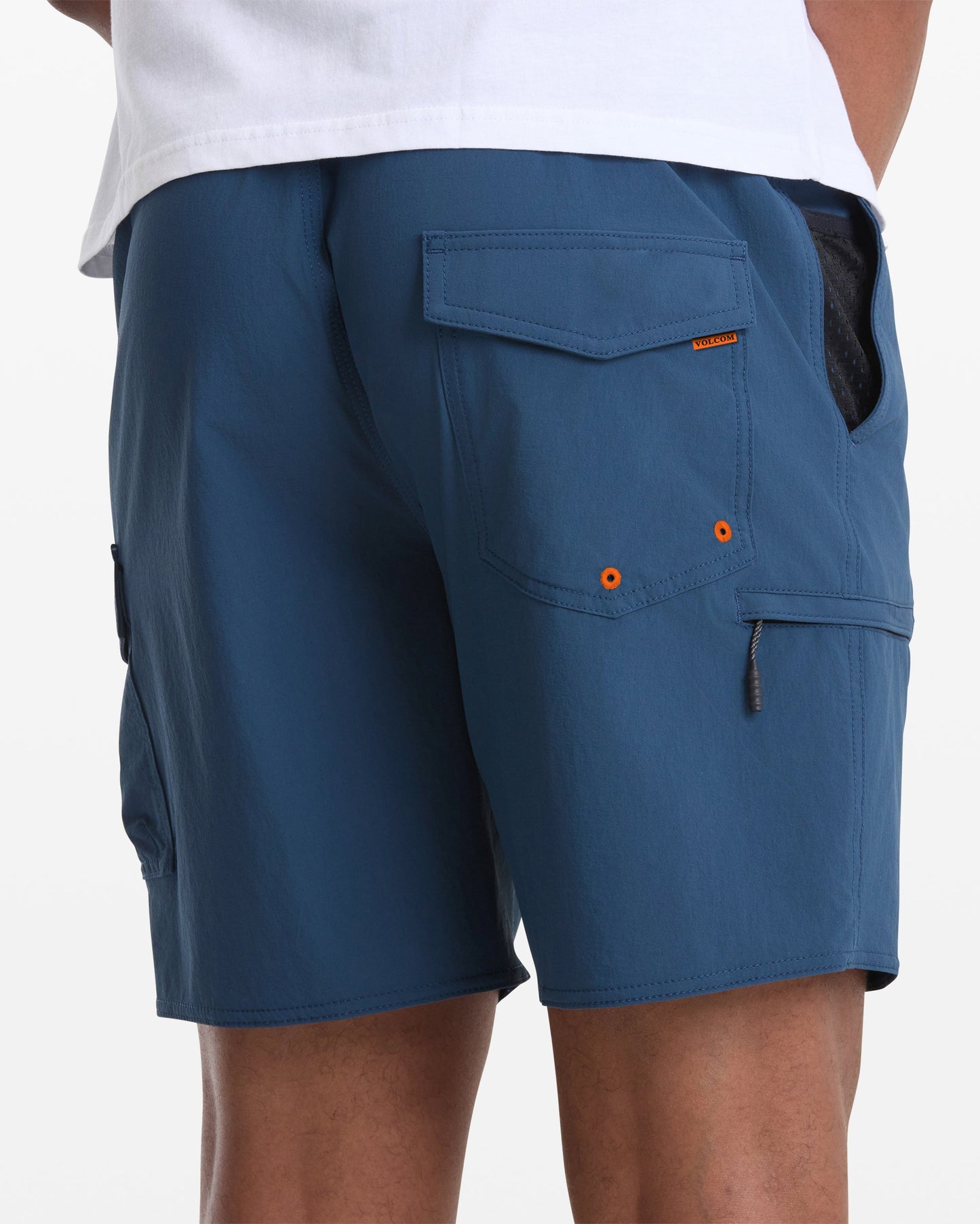 A person wearing Volcom Men's Truly Lib Work Trunk 17 blue cargo shorts with flap pockets and orange accents stands with their back to the camera, showing the lower part of a white shirt.