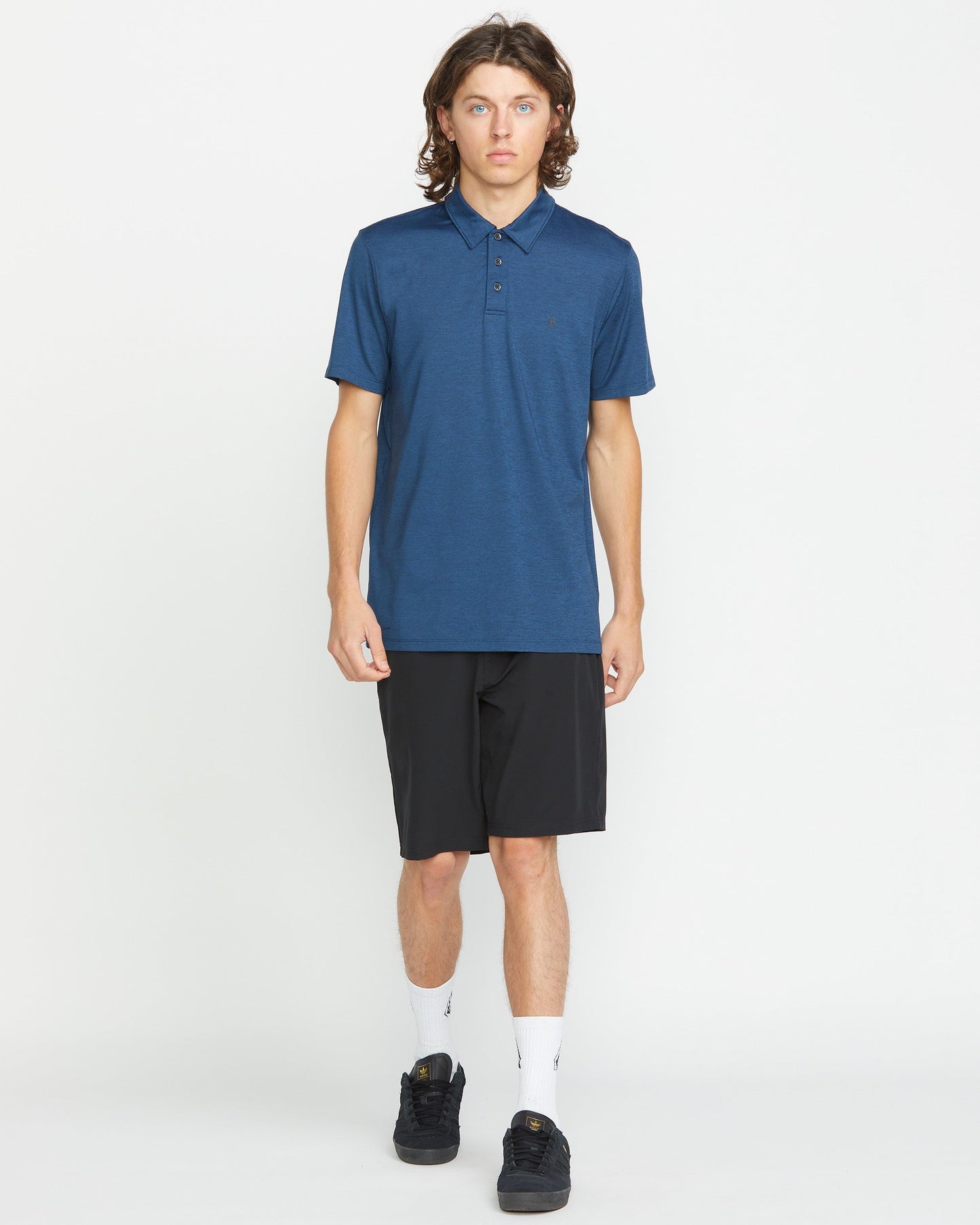 A young man with long brown hair wears a blue polo, Volcom Men's Frickin Cross Shred 20" Shorts, white socks, and black sneakers while standing against a plain white background.