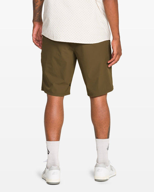 Only the lower half of a person is visible, wearing an off-white shirt, white socks and sneakers, and Volcom Men's Frickin Cross Shred Shorts in water-repellent olive green with a 20" outseam. Their back faces the camera.