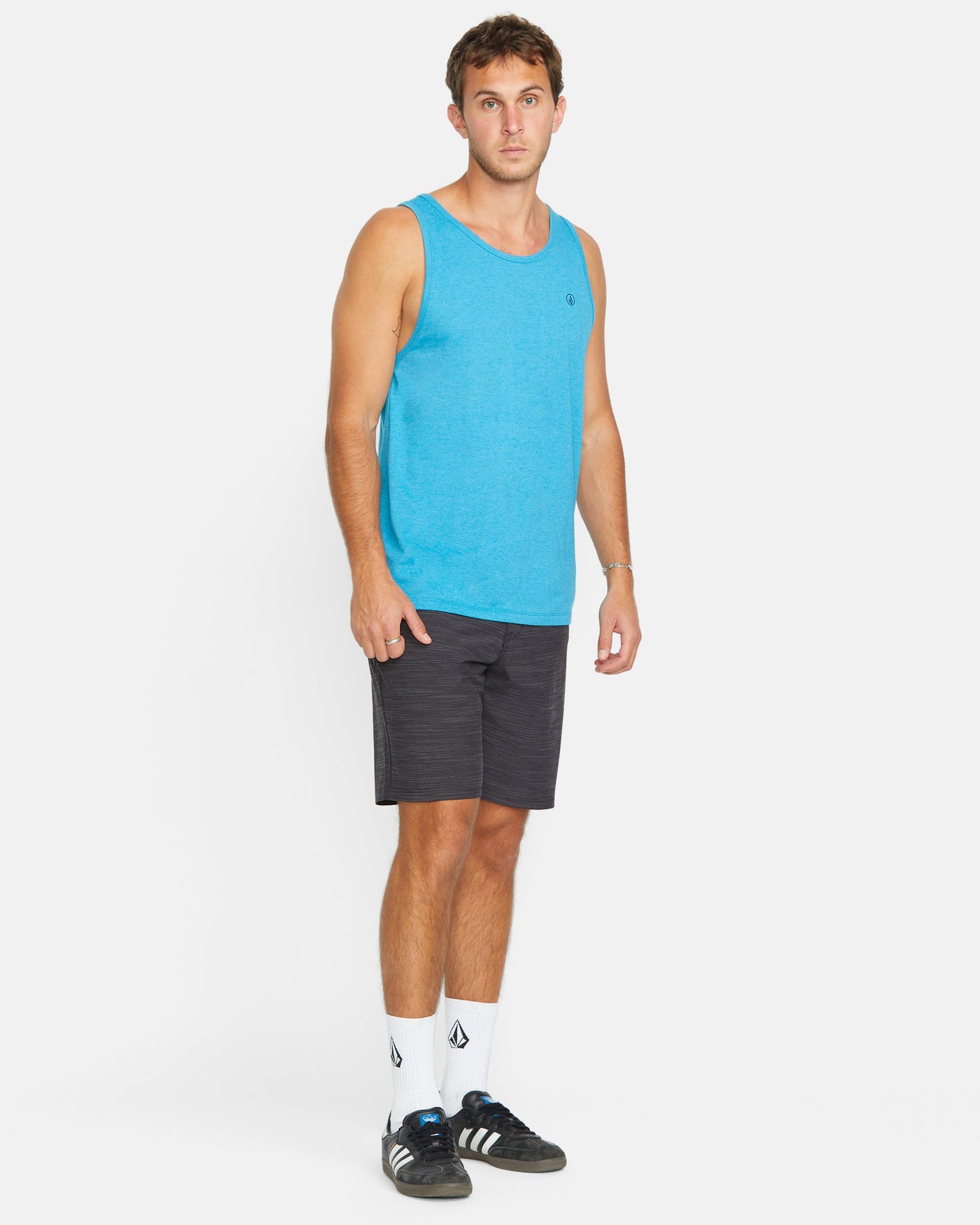 A man faces forward, wearing a bright blue tank top, Volcom Men's Slub Frickin Cross Shred 20" Hybrid Shorts, white crew socks with a black logo, and black athletic shoes against a plain white background.