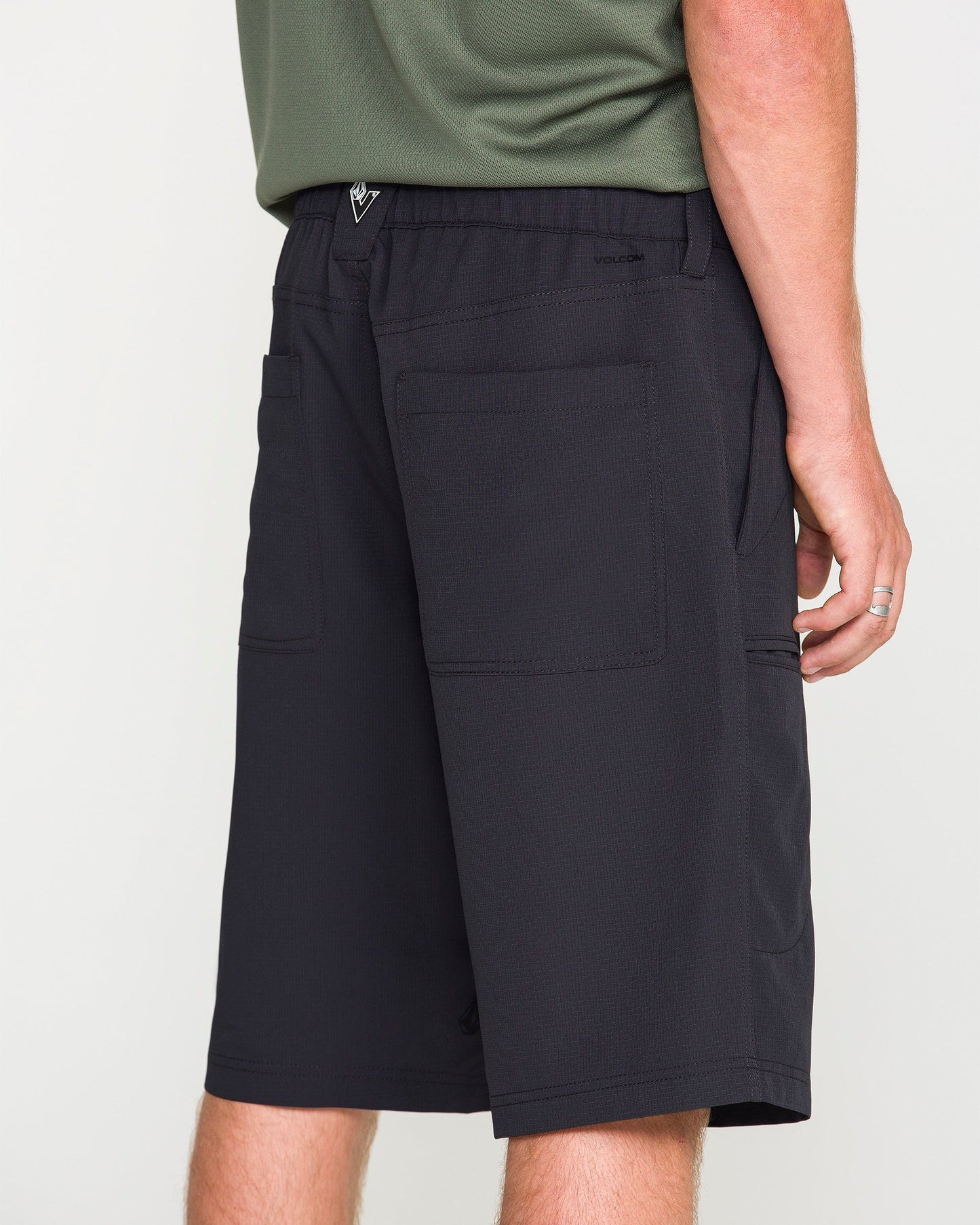 A person wearing Volcom Men's Venture Stone Hybrid Shorts in Asphalt Black, featuring knee-length recycled-poly fabric and large back pockets, along with a green shirt and a silver ring, standing against a plain white background.