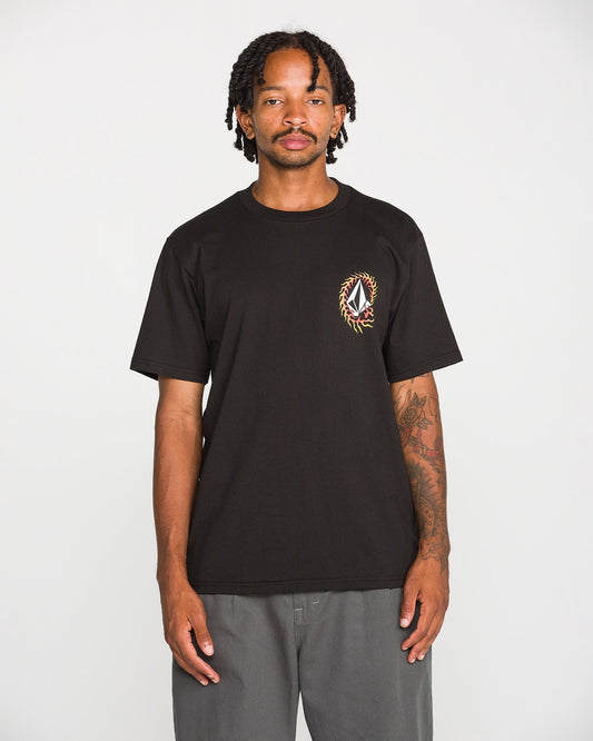 A person with shoulder-length dreadlocks wears the Volcom Men's Ringafire Short Sleeve Tee in classic black, crafted from heavy-weight premium cotton and featuring a small, colorful crest on the left chest. They pair it with gray pants and stand against a plain white background.
