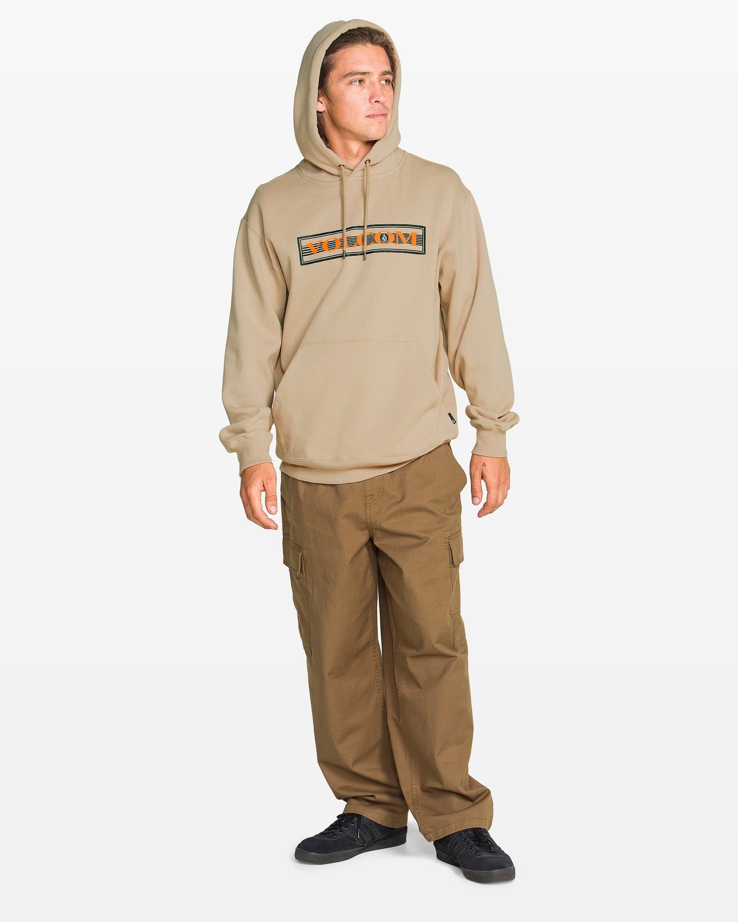 A man wears a tan Volcom Men's Stone Coast Pullover, brown cargo pants, and black shoes while standing slightly to the side against a plain white background.