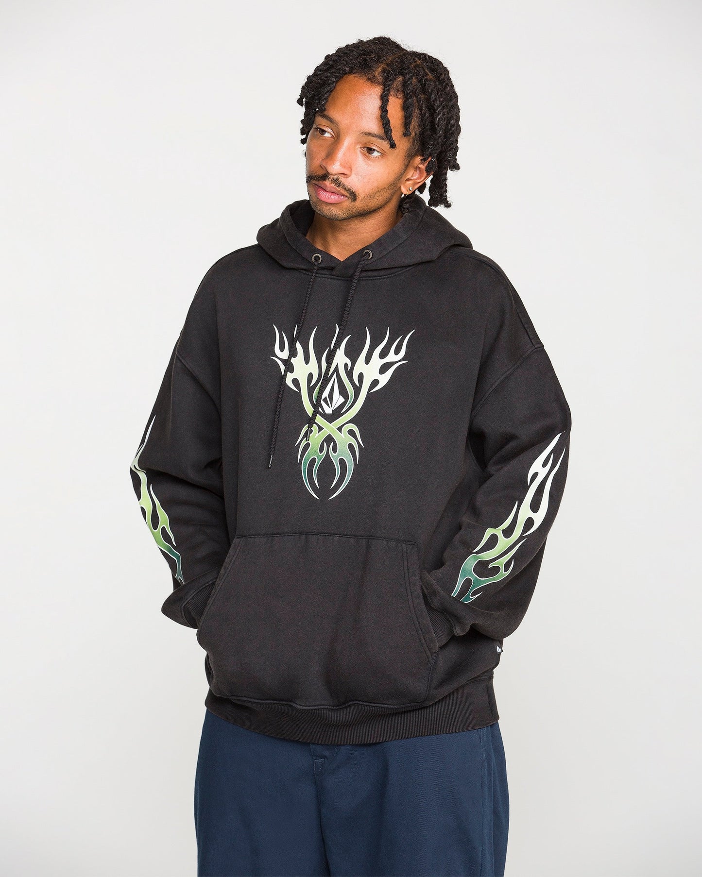 A person wearing the Volcom Men's Stone Trip Pullover—a washed-black hoodie featuring white and green flame designs on the chest and sleeves—stands with hands in the front pocket, looking slightly to the side.