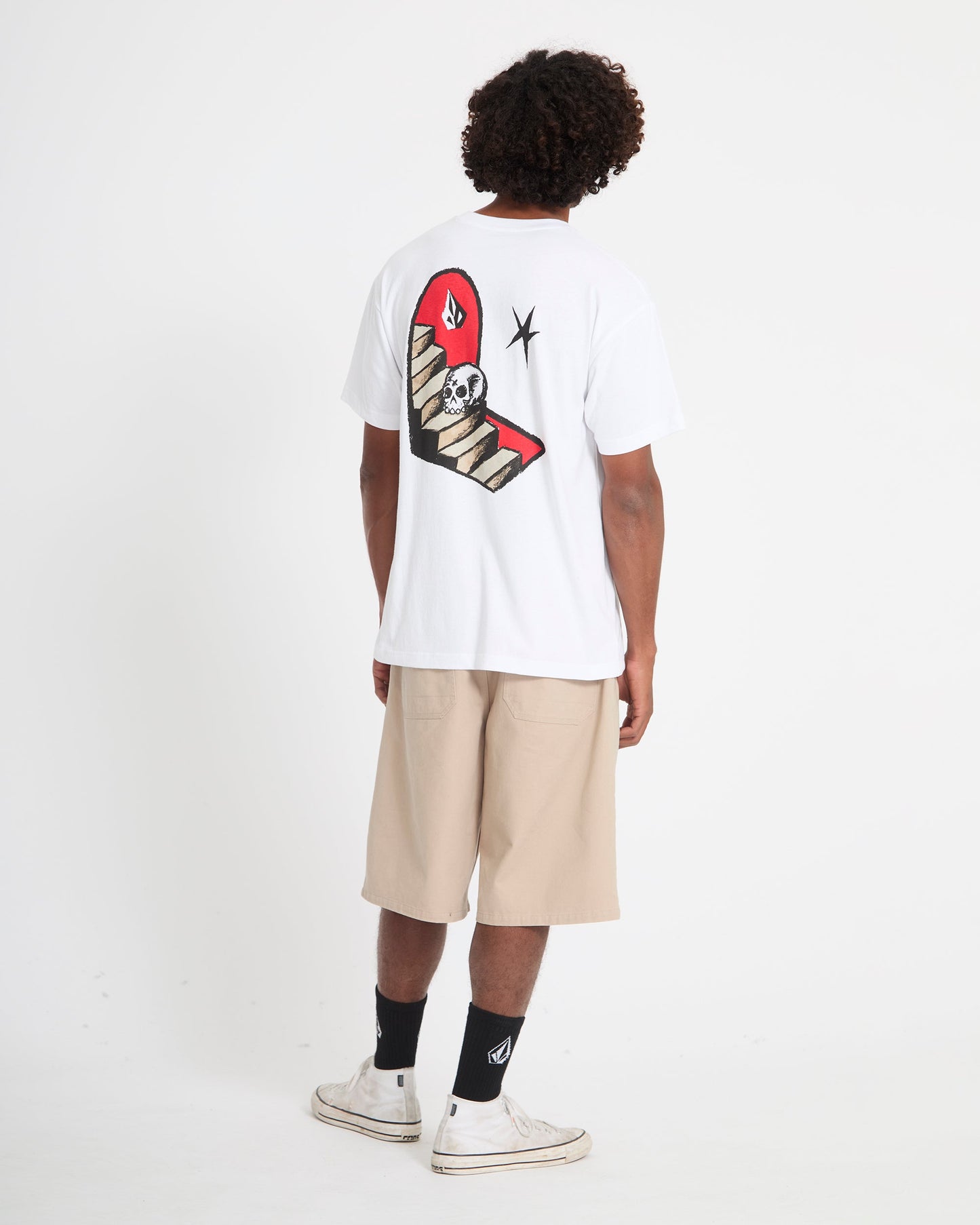 A person models the Volcom Men's Featured Artist Seth Conboy Loose Short Sleeve Tee, which has a large graphic on the back. They're also wearing beige shorts, black socks, and white sneakers against a plain white background.