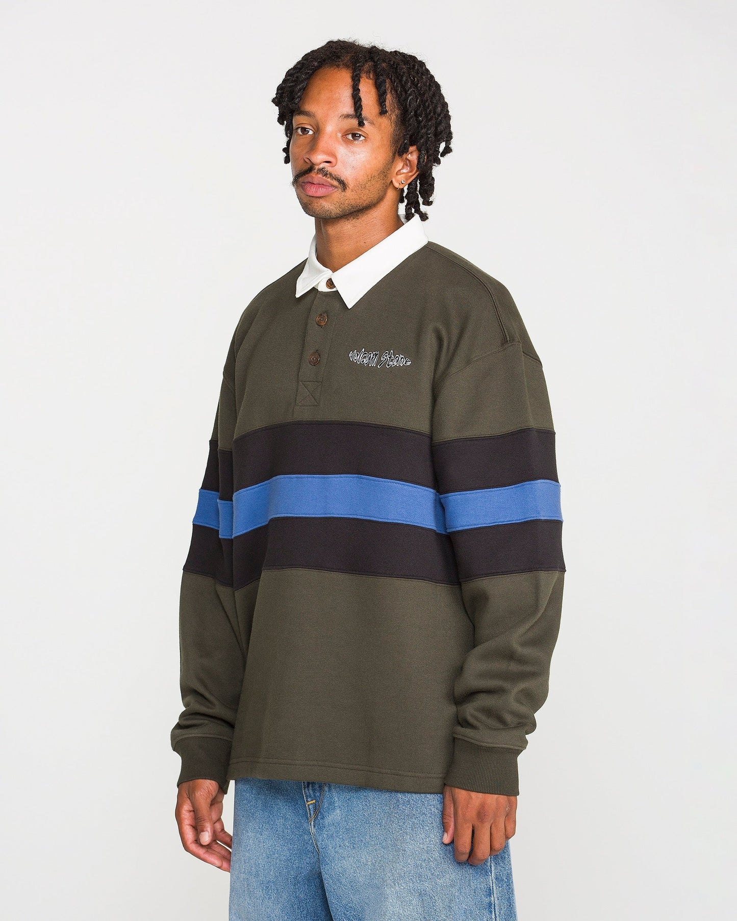 A person wearing the Volcom Men's Scratch Stone Fleece Polo—a dark green long-sleeve shirt with black and blue horizontal stripes in a boxy fit—and light blue jeans stands against a plain white background.