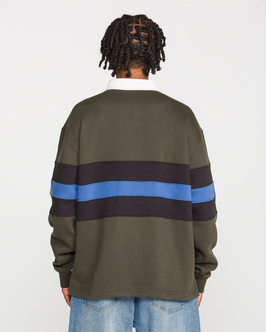 A person with short twists stands facing away, wearing the Volcom Men's Scratch Stone Fleece Polo in a boxy fit olive green with black and blue horizontal stripes, crafted from recycled polyester fleece, paired with light blue jeans against a plain white background.