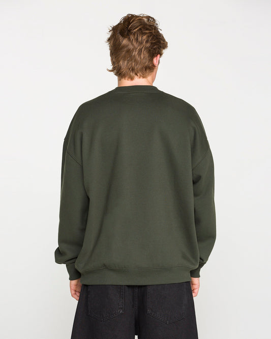 A person with short, light brown hair is facing away from the camera, wearing the Volcom Men's Dirty Work Crew—an oversized green sweatshirt crafted from recycled polyester—paired with black pants against a plain white background.