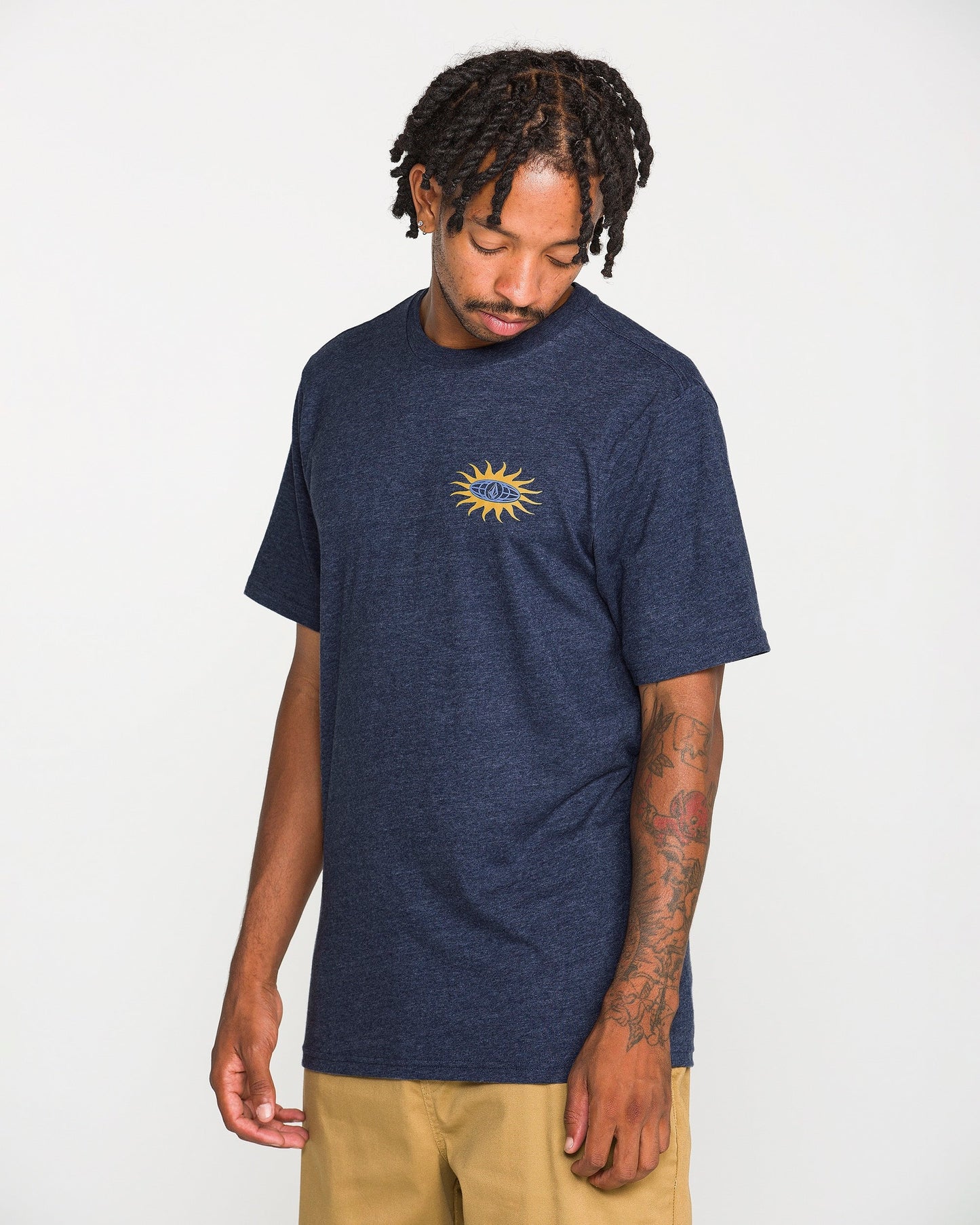 A man stands against a plain white background, looking down with his right hand by his side. He wears tan pants and the Volcom Men's Spf91 Short Sleeve Tee—a navy blue, medium-weight heather shirt featuring a yellow sun graphic on the chest. His left arm is adorned with tattoos, completing the perfect laid-back look in this classic tee.