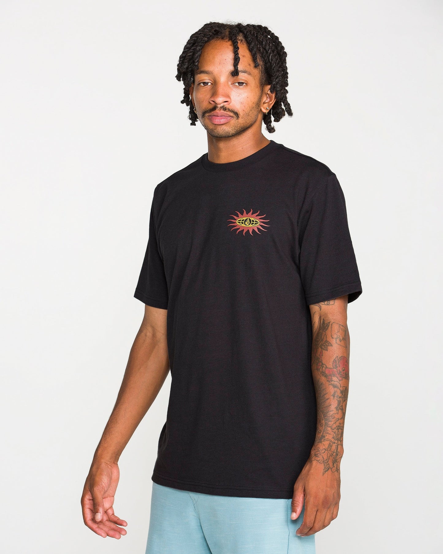 A man with short dreadlocks is wearing the Volcom Men's Spf91 Short Sleeve Tee in a black, medium-weight heather. The shirt features a small sun and eye graphic on the chest and is designed with a modern fit, crafted from a soft ring spun cotton-poly blend for everyday comfort. He stands against a plain white background.