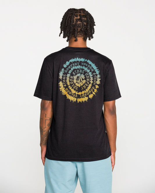 A person with short dreadlocks wears the Volcom Men's Still Spiraling Short Sleeve Tee, a washed black t-shirt featuring a colorful circular graphic and wavy text created with a soft hand screen print on the back. They pair it with light blue shorts while standing against a plain white background.