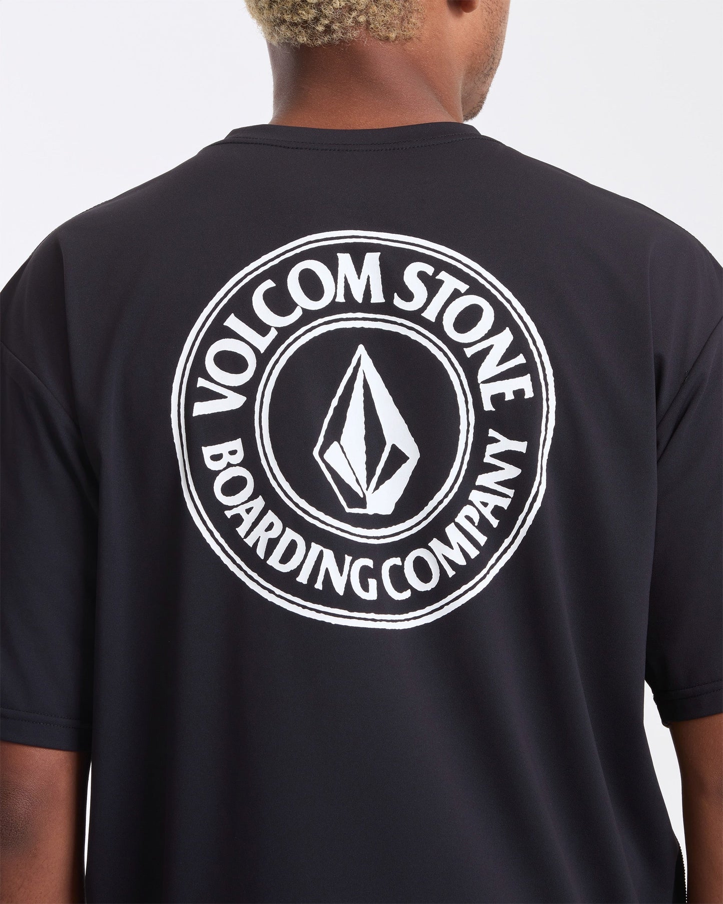 A man wearing the Volcom Men's Drain You Short Sleeve Tee, featuring a large white circular Volcom Stone Boarding Company logo and diamond emblem on the back—perfect for aquatic adventures.