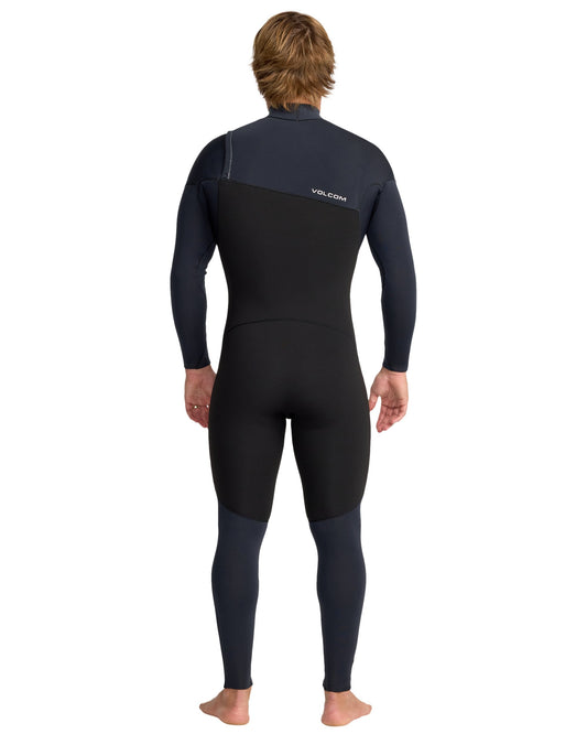 A person with light brown hair, barefoot and facing away, wears the Volcom Men's 4/3 Modulator Chest Zip Full Wetsuit in black and dark navy, featuring long sleeves and the Volcom logo on the upper back.