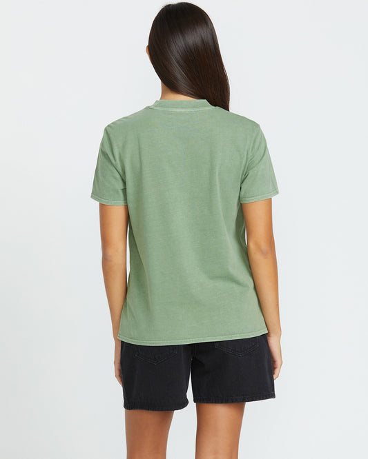 A woman with long dark hair, seen from behind, wears the Volcom Women's Lock It Up Tee in light green with black denim shorts. Her relaxed style stands out against the plain white background.