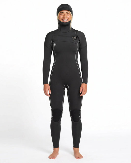 A person stands barefoot and smiling against a plain white background, wearing the Volcom Women's 4/3MM Modulator Chest Zip Hooded Full Wetsuit with white seams.