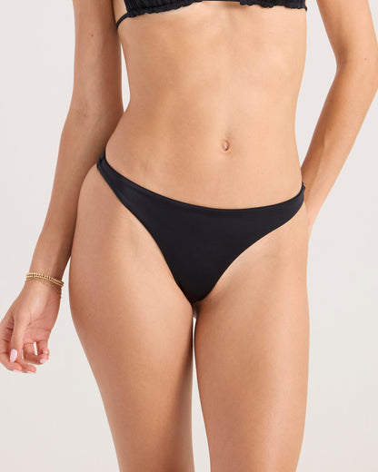 A model wears the Volcom Women's Simply Seamless Scoop Tiny black bikini bottom and matching top, shown from mid-torso to upper thighs against a plain background. Made from recycled nylon for an eco-friendly touch.