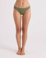 A woman stands barefoot against a plain background, wearing Volcom Women's Simply Seamless Hipster Full bikini bottoms in olive green. Her arms are out of frame, and her body is facing forward, showcasing the comfortable full coverage swimwear design.