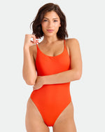 A woman with shoulder-length brown hair wears the Volcom Women's Simply Seamless Skimpy One-Piece swimsuit in orange, standing against a plain light background and looking at the camera.