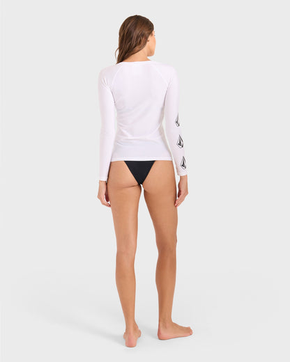 A woman stands barefoot facing away, wearing the Volcom Women's Simply Core Long Sleeve Rashguard with black logos on the left sleeve and black bikini bottoms. The background is plain light gray.