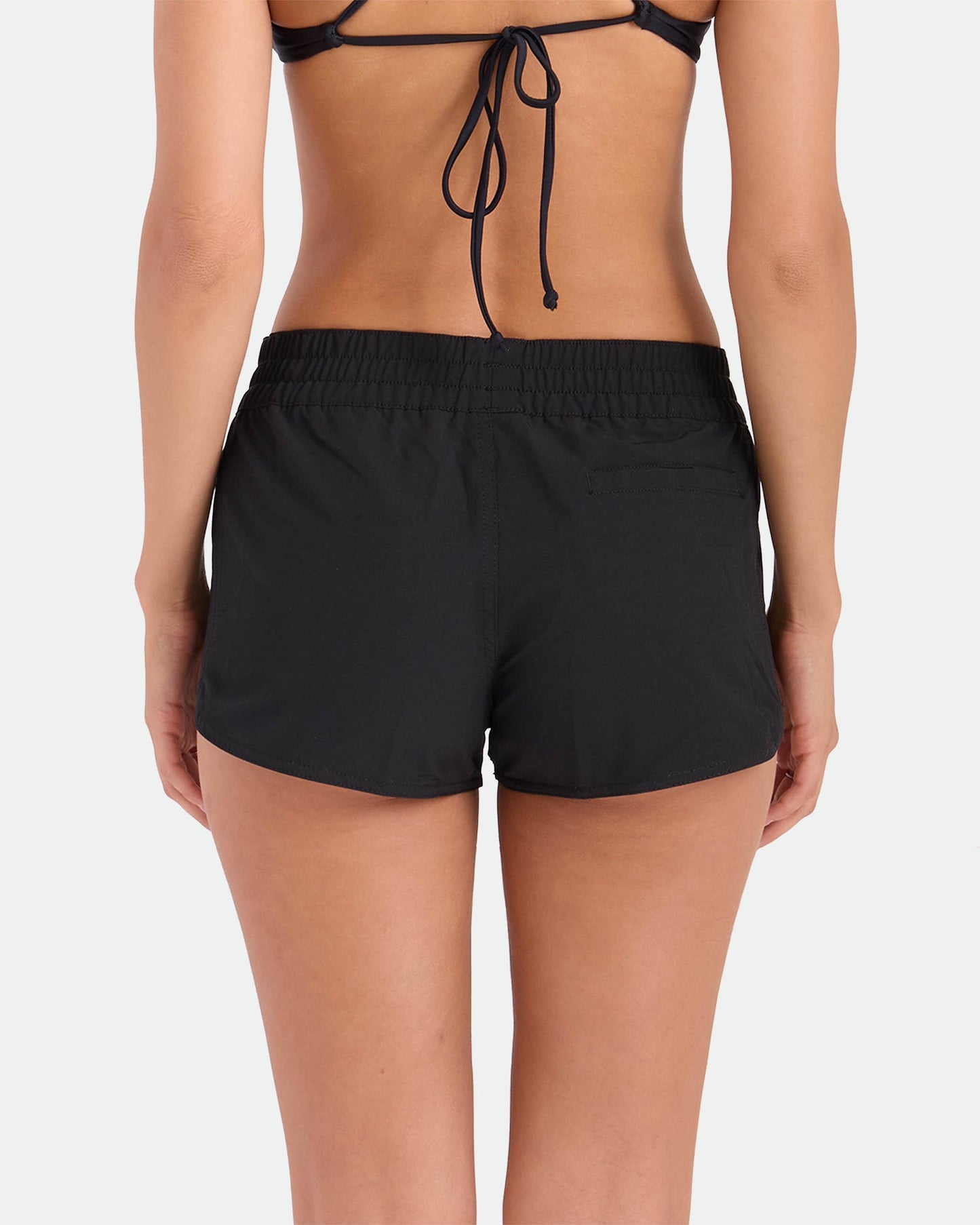 A woman is seen from the back in a black bikini top and Volcom Women's Simply Solid Boardshorts with an elastic waistband, standing against a plain white background.