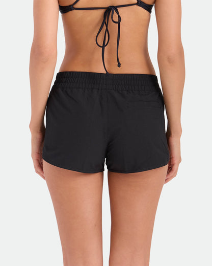A woman is seen from the back in a black bikini top and Volcom Women's Simply Solid Boardshorts with an elastic waistband, standing against a plain white background.