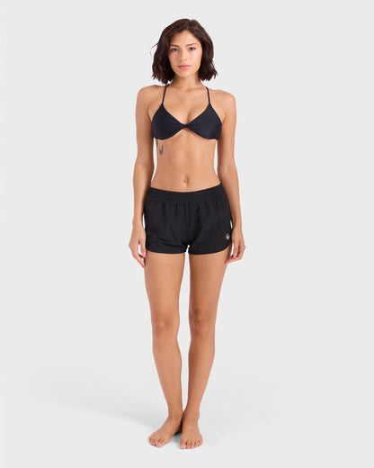 A woman stands barefoot against a plain light background, facing forward with arms relaxed by her sides, wearing a black bikini top and Volcom Women's Simply Solid Boardshort made from recycled polyester.