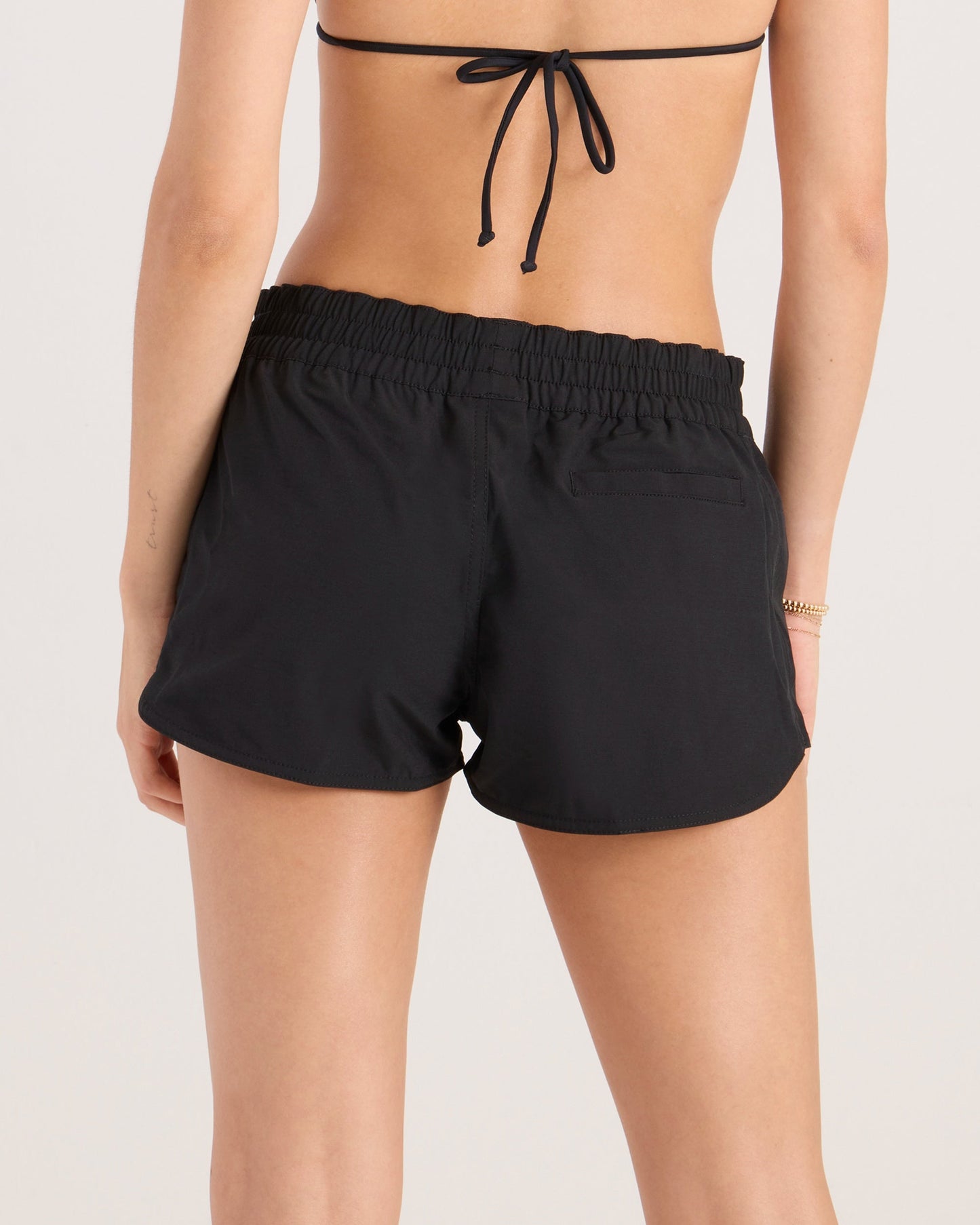 A woman is shown from the back wearing a black bikini top and Volcom Women's Simply Solid Boardshorts, made from recycled polyester with 4-way stretch, elastic waistband, UPF 50+ protection, and a small back pocket.