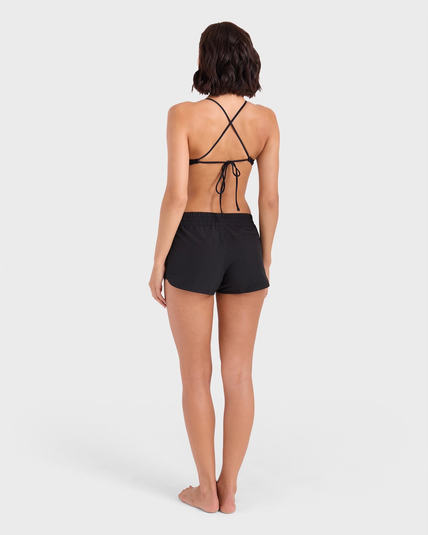 A woman with wavy, shoulder-length hair stands barefoot facing away, wearing the Volcom Women's Simply Solid Boardshort and a black strappy bikini top against a plain white background.